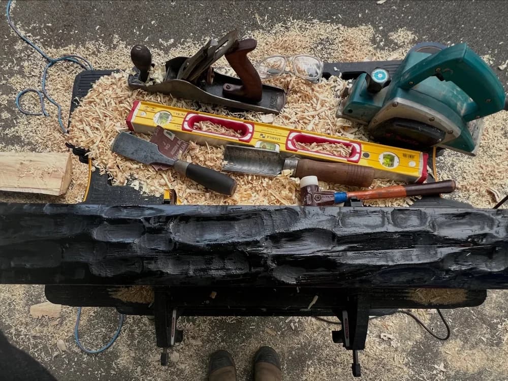 A blackened hand-hewn timber mantel on sawhorses surrounded by hand tools, a level and wood shavings