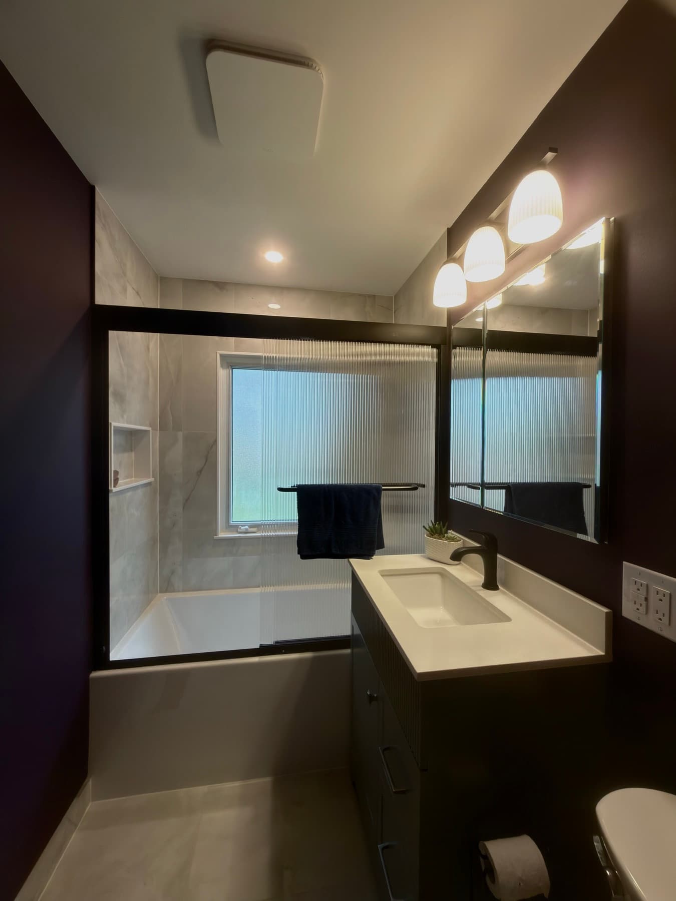 Reeded glass tub enclosure beside a dark vanity in a deep plum bathroom