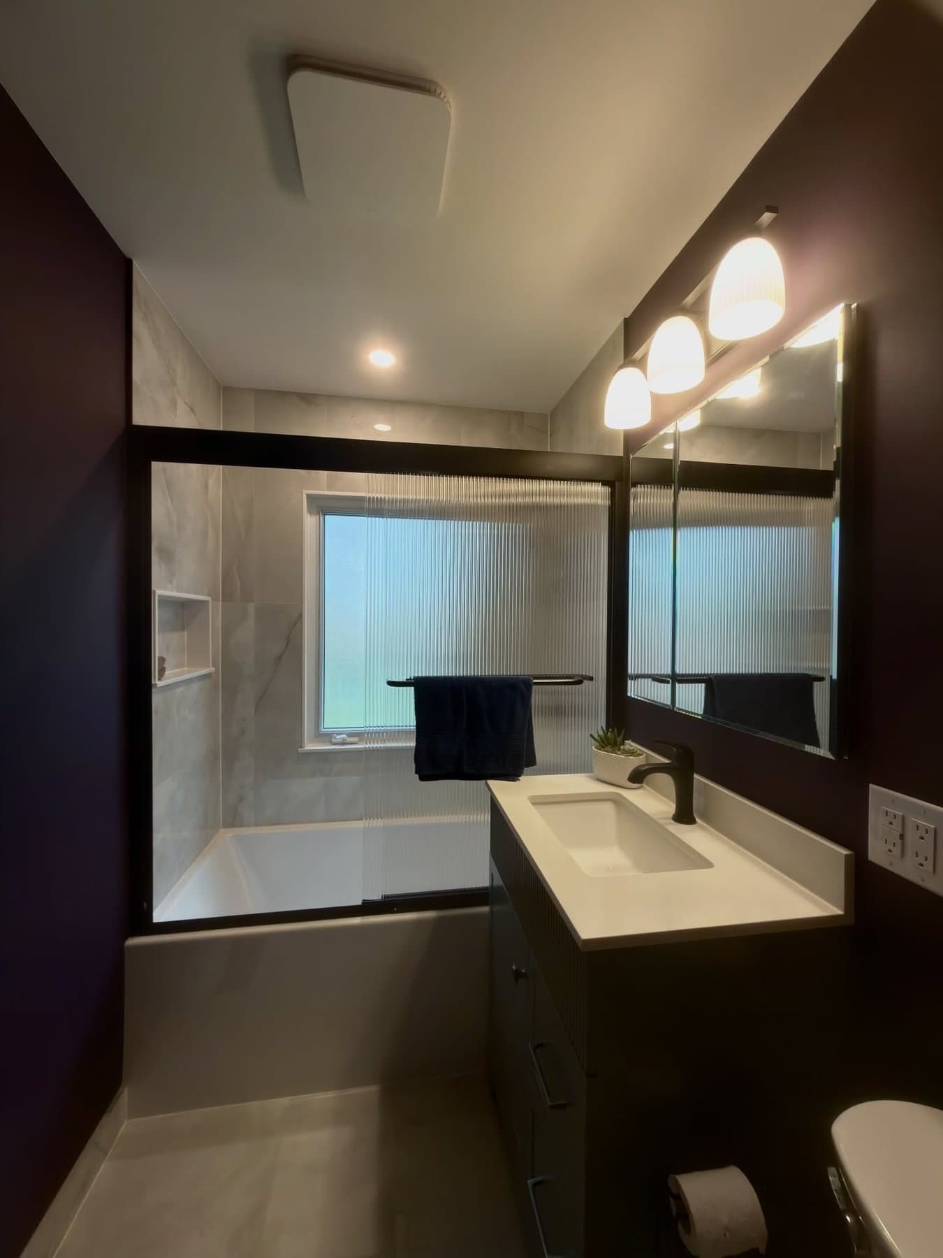 Wide view of the plum bathroom showing the fluted vanity, reeded glass tub enclosure and tiled niche together