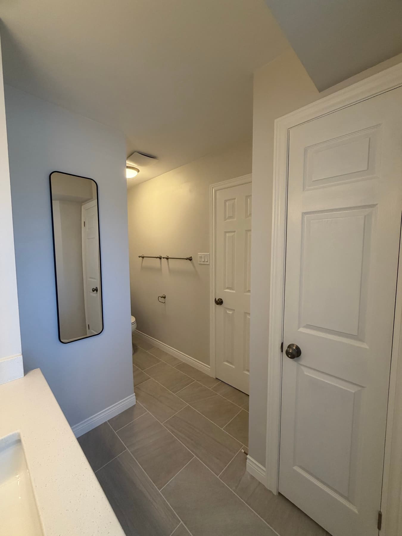Tall mirror on a grey wall beside a hallway with warm grey plank tile leading to two doors