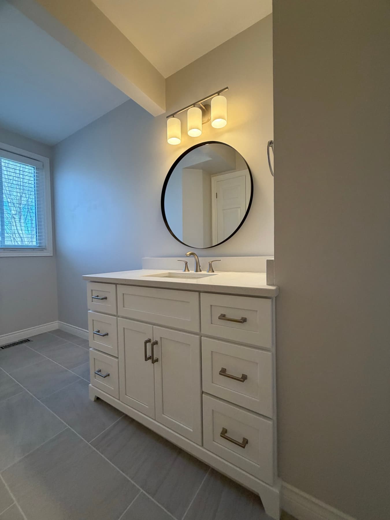 Bright bathroom vanity area with a round black-framed mirror over a white shaker vanity and grey plank tile floor