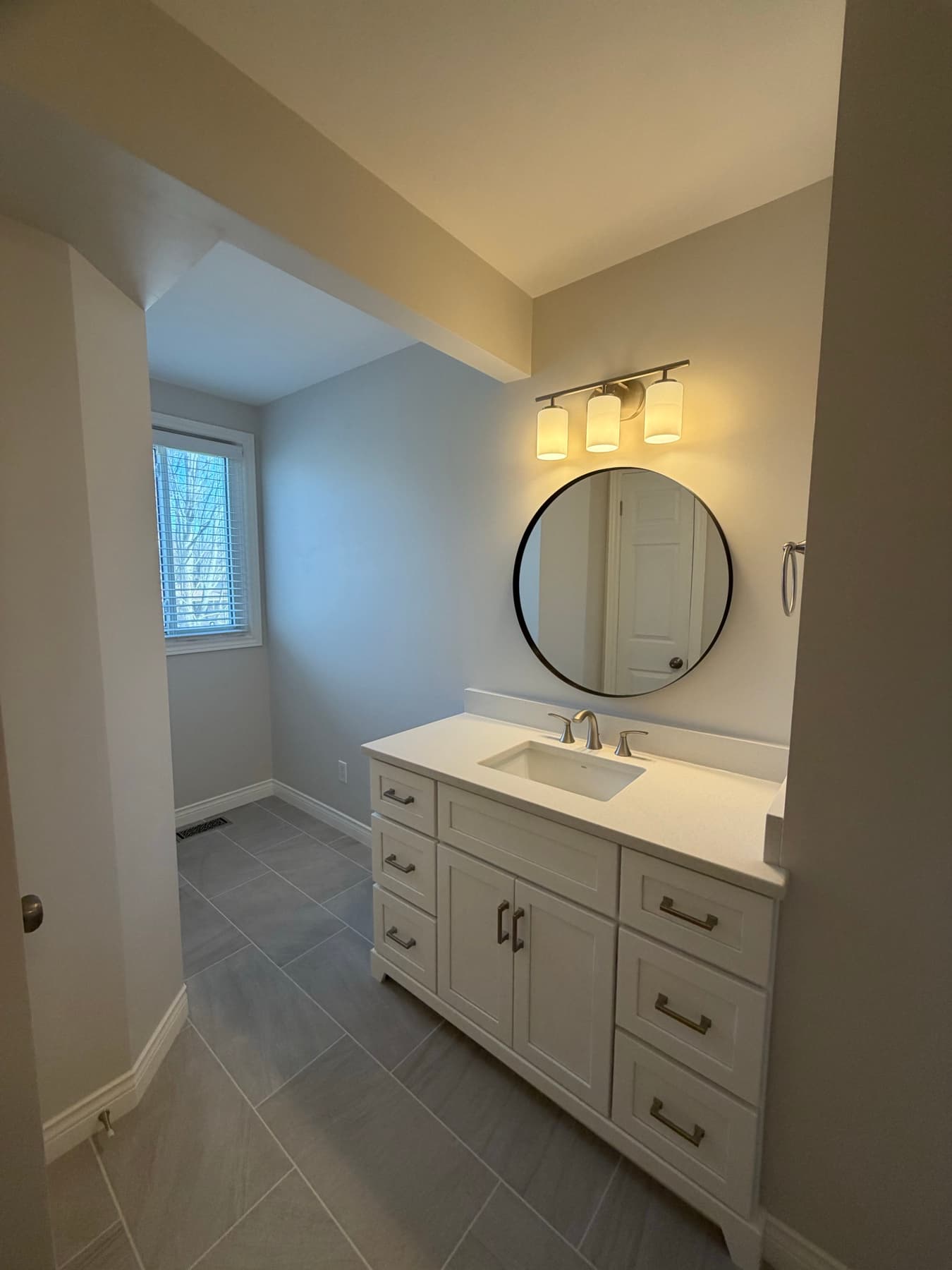 White shaker vanity with a round mirror, seen from the adjoining hallway with grey plank tile underfoot