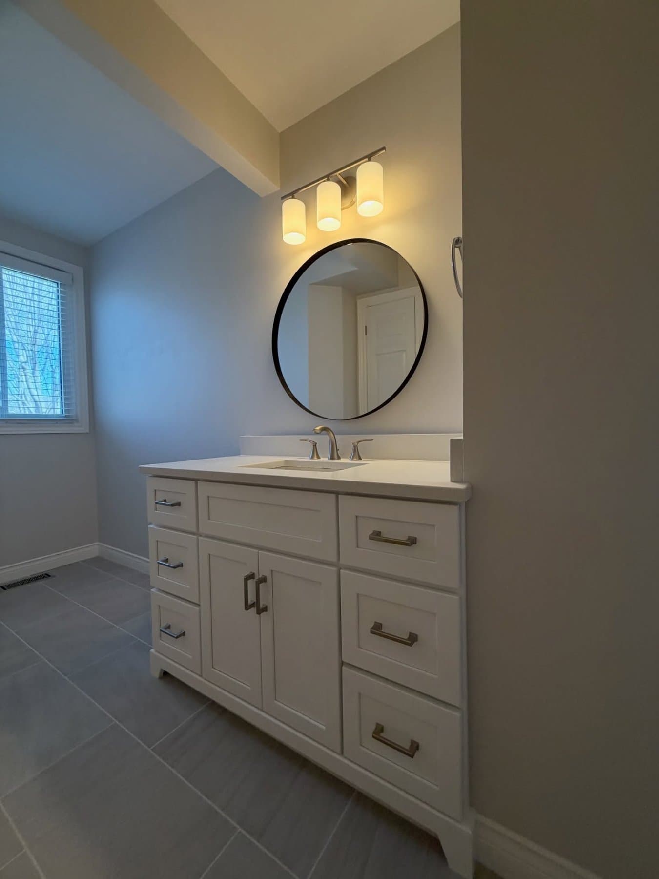 White shaker vanity seen from across the room with a window and round mirror, grey plank tile underfoot