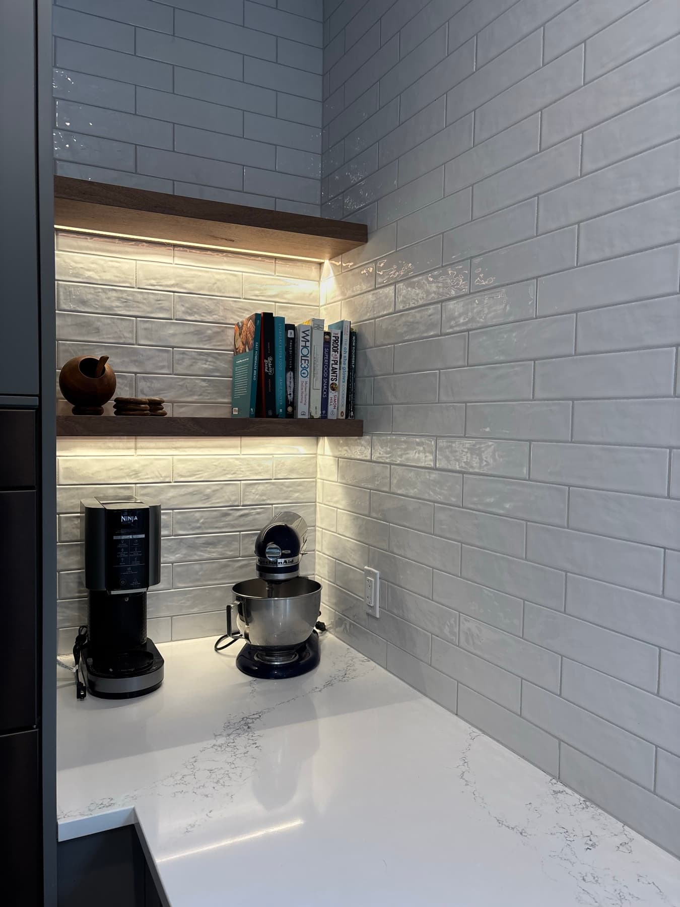 Floating wood shelves over a coffee station, backed by white tile with integrated lighting