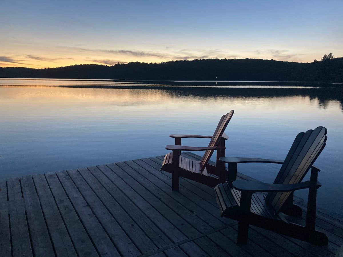 Two Muskoka chairs on a wooden dock overlooking a calm lake at sunset