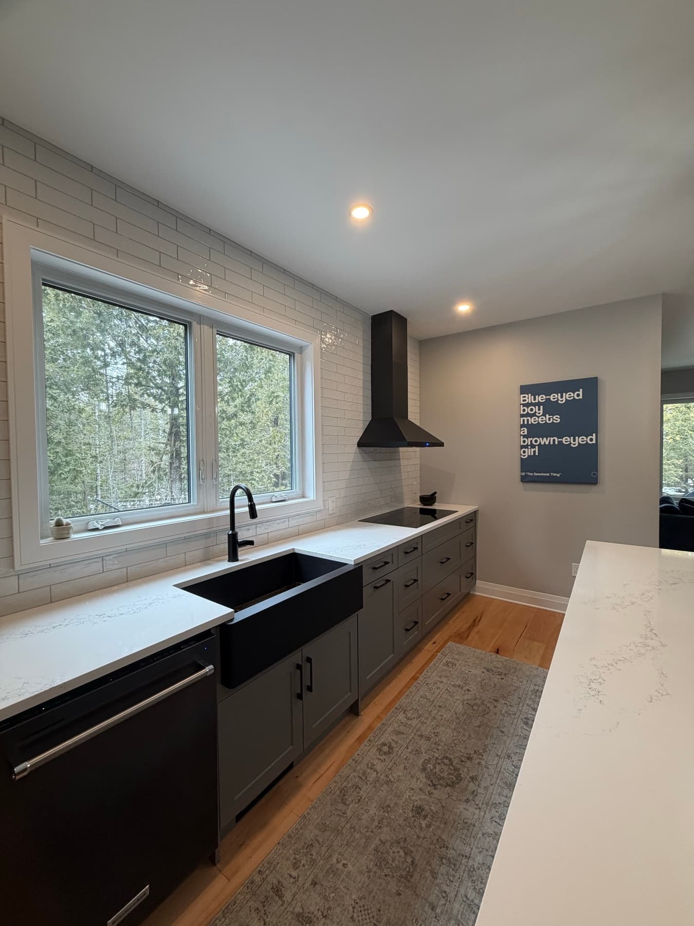 A black farmhouse sink under a large window looking into the trees, with black shaker cabinetry and a black range hood